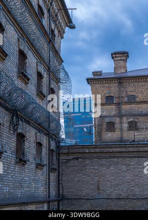 La prigione di Lukiskes a Vilnius, in Lituania, mostra finestre sbarrate, filo di rasoio e luci di sicurezza, con un alto edificio di vetro blu dietro sotto un freddo coperto Foto Stock
