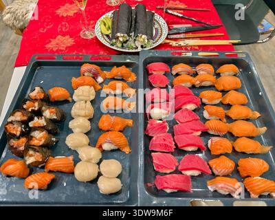 Sushi fatto in casa preparato per gli ospiti a una festa. I sushi in primo piano sono su vassoi culinari. Sullo sfondo sono presenti rotoli su una piastra su un tavolo con un rosso Foto Stock