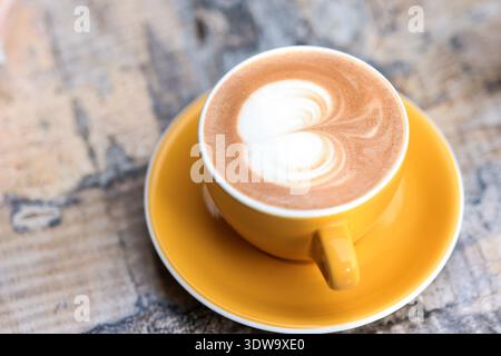 Cappuccino caldo con latte Art su tazza gialla sopra il tavolo rustico Foto Stock