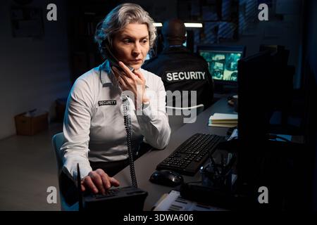 Donna caucasica di mezza età che monitora le telecamere di sicurezza e parla al telefono nella sala di controllo, seduto alla scrivania con il computer mentre l'agente di sicurezza Black Man lavora in background Foto Stock