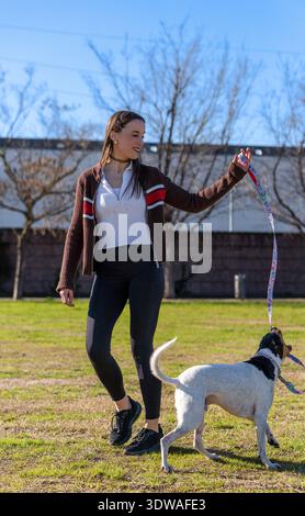 Una bella e atletica giovane donna che indossa un cardigan marrone e leggings neri gioca con il suo cane maculato su un guinzaglio colorato in un parco soleggiato Foto Stock