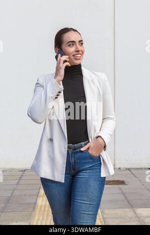 Una donna cammina su un marciapiede mentre utilizza uno smartphone. È vestita con un blazer e un jeans. Questa scena si svolge in un'area urbana durante il giorno Foto Stock