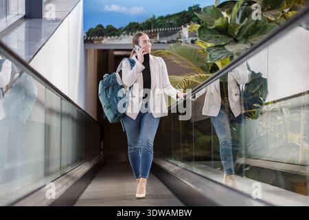 Una donna sta camminando attraverso un moderno corridoio di vetro, usando il suo telefono mentre indossa uno zaino. La scena la cattura in un ambiente luminoso, suggesti Foto Stock