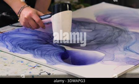 La foto ravvicinata mostra un artista che asciuga gli strati di inchiostro su tela con uno strumento di riscaldamento, una luce soffusa per interni e un tavolo creativo disordinato. Fare arte supporta le emozioni Foto Stock