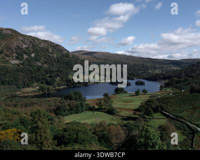 Vista aerea della tranquilla Rydal Water, annidata tra colline verdi ondulate e foreste lussureggianti sotto un cielo luminoso, Ambleside, Inghilterra, Regno Unito. Foto Stock