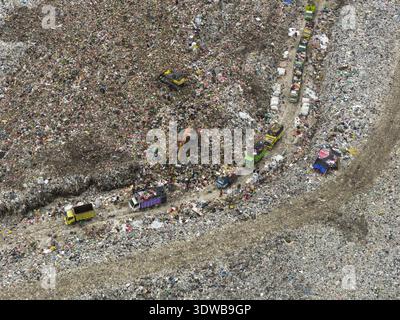Vista aerea di una vasta discarica, un paesaggio aspro di rifiuti attraversato da una processione di camion colorati, in netto contrasto con la natura circostante, Foto Stock
