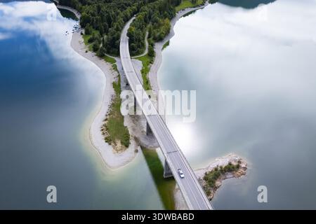 Vista aerea di un ponte che attraversa il tranquillo lago che rispecchia il cielo, collegando lussureggianti masse verdi, Sylvensteinspeicher, Bayern, Germania. Foto Stock
