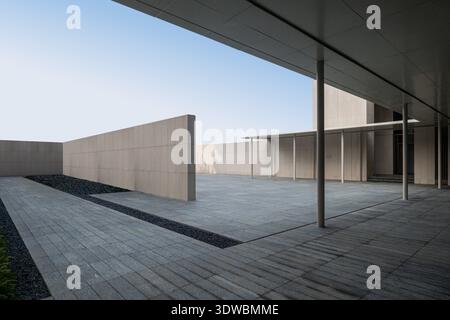 Un'ampia prospettiva architettonica di un cortile minimalista vuoto con pavimento in piastrelle di pietra e pareti geometriche in cemento sotto un cielo limpido. Foto Stock