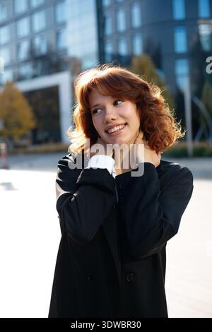 Allegra giovane donna d'affari sorridente con una posa rilassata e toccante sul collo in piena luce del sole della città. Foto Stock