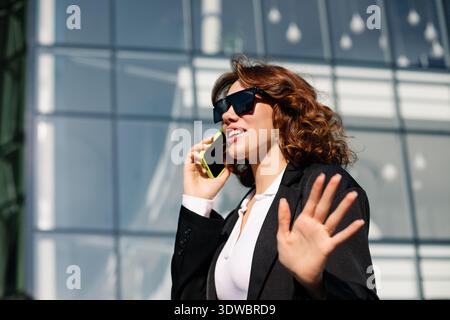 Donna d'affari sicura che cammina all'aperto mentre parla su smartphone, stile di vita aziendale dinamico e concetto di comunicazione. Foto Stock