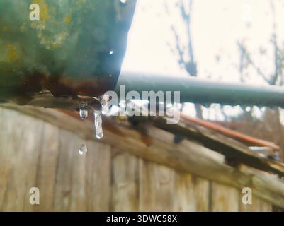 Una foto macro ad alta risoluzione che cattura la delicata bellezza della fusione del ghiaccio. Caratterizzato da ghiaccioli semitrasparenti e una goccia d'acqua a mezz'aria da un tetto arrugginito Foto Stock
