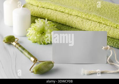 Carta per la cura della pelle con rullo facciale in giada per massaggio anti-invecchiamento della pelle, fiori e asciugamani morbidi su un tavolo di marmo. Foto Stock