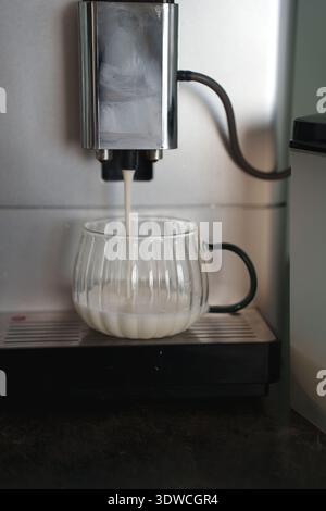 macchina per il caffè automatica che versa il latte montato in una tazza di vetro trasparente su un bancone della cucina con accenti neri e in acciaio inox sotto la luce naturale Foto Stock