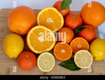 Un primo piano ad alto angolo di agrumi misti con arance, limoni e mandarini a fette e intere con foglie verdi su un tagliere di bambù. T Foto Stock
