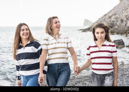 Una scena gioiosa di una madre e delle sue due figlie che camminano lungo la spiaggia, tenendosi per mano e sorridendo. Le onde oceaniche si infrangono dolcemente sulla riva, creando un Foto Stock
