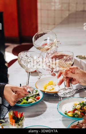 Ritagliato irriconoscibile. Tre mani preparano un brindisi celebrativo con bicchieri da vino, spruzzando del liquido sopra un tavolo con vivaci insalate e colorate di Foto Stock