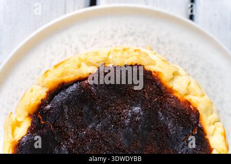 Una deliziosa cheesecake basca si trova su un rustico piatto bianco, caratterizzato dalla sua caratteristica cima bruciata e da una cremosa all'interno. Il dessert è iconico per il suo carattere unico Foto Stock