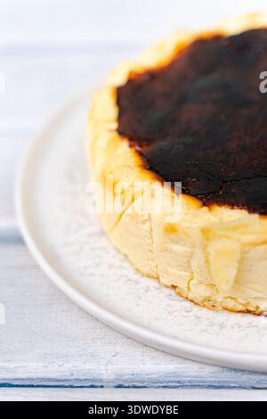 Una deliziosa cheesecake basca con un top perfettamente tostato si siede su un piatto bianco su un rustico tavolo di legno, mostrando la sua consistenza cremosa e Foto Stock