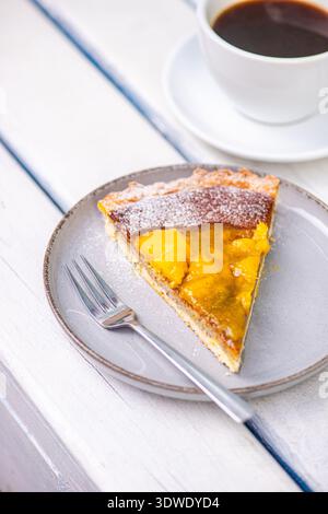 Una deliziosa fetta di torta al mango si trova su un piatto grigio con una forchetta, accompagnata da una tazza di caffè nero fumante, perfetto per un dolce finale Foto Stock