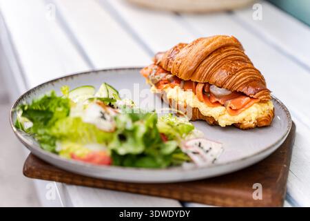 Un croissant appena sfornato ripieno di salmone affumicato e uova cremose, servito insieme a una vivace insalata verde, per creare un pasto delizioso e salutare Foto Stock