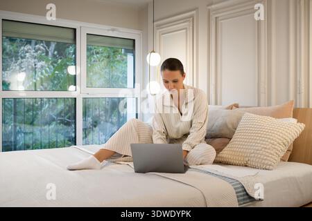 Una giovane donna in comode poltrone siede su un letto in una stanza accogliente e ben illuminata, coinvolgendo il suo laptop. L'atmosfera e' tranquilla e moderna, sugge Foto Stock
