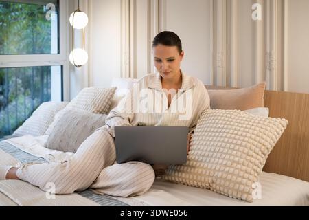 Una giovane donna che indossa un comodo pigiama sta sdraiandosi su un comodo letto con cuscini, coinvolgendo il suo laptop. Un'illuminazione soffusa crea un ambiente tranquillo Foto Stock