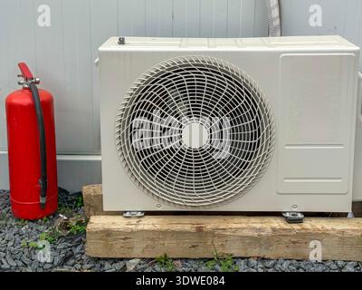 Condizionatore d'aria esterno posizionato su supporti in legno, accompagnato da un estintore rosso. La configurazione mette in evidenza sicurezza e funzionalità in un ou Foto Stock