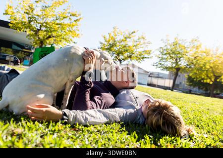 I fratelli si godono un momento di gioia all'aperto, sdraiati sull'erba sotto il sole, giocando con il loro cane amico, creando una scena di amicizia e. Foto Stock