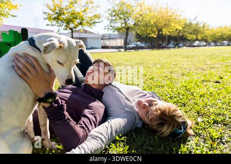 I fratelli si godono un momento di gioia all'aperto, sdraiati sull'erba sotto il sole, giocando con il loro cane amico, creando una scena di amicizia e. Foto Stock