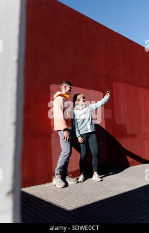 I fratelli si godranno una giornata d'autunno soleggiata in città, scattando un selfie contro un'audace parete urbana rossa. Il loro abbigliamento casual e le pose allegre riflettono un vibr Foto Stock