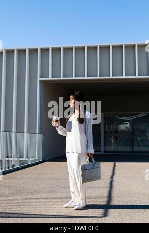 Una donna professionale si trova all'esterno di un edificio moderno, gustando un caffè alla luce del sole mattutino. È vestita di bianco e porta un'elegante ha grigia Foto Stock
