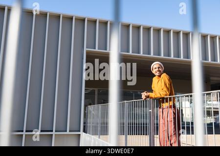 Un uomo sta accanto a una ringhiera all'aperto, sorridendo calorosamente mentre indossa un abbigliamento casual con un maglione arancione e un cappello. L'architettura moderna fornisce un Foto Stock