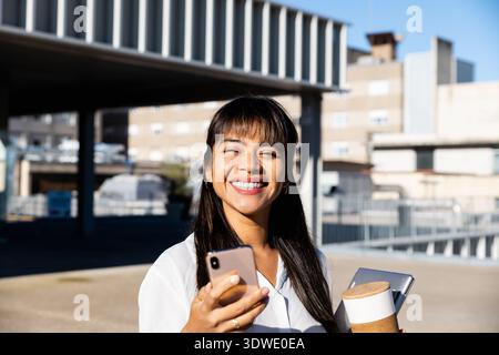 Una donna d'affari sicura di sé sta all'aperto tenendo in mano uno smartphone e una tazza di caffè, sorridendo brillantemente in un ambiente professionale, mostrando il business moderno Foto Stock
