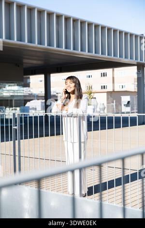 Una donna sta all'aperto, sorridendo mentre parla al telefono. Si appoggia casualmente contro una ringhiera, bagnata dalla luce del sole, con edifici moderni nel bac Foto Stock