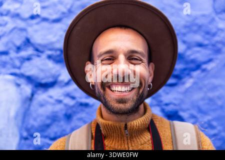 Un uomo sorridente di fronte a un muro blu vibrante a Chefchaouen, in Marocco. Il dettaglio del suo cappello marrone e del maglione senape aggiunge calore e contrasto al Foto Stock