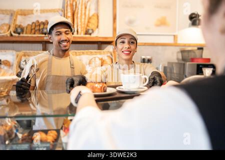 I lavoratori sorridenti del caffè servono pasticcini freschi e caffè a un cliente in un accogliente ambiente da forno. Il pane viene visualizzato sullo sfondo, creando un welcom Foto Stock