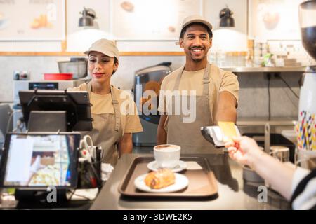 Due simpatici lavoratori del caffè in uniforme servono un cliente in una panetteria moderna. Un caffè e una pasticceria su un vassoio indicano l'atmosfera accogliente e la qualità Foto Stock