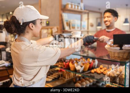 Un addetto al bar serve un cliente sorridente al bancone in un'accogliente panetteria. Il espositore è pieno di una varietà di deliziosi pasticcini e muffin Foto Stock