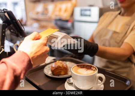Un cliente effettua un pagamento contactless presso un accogliente bancone di una panetteria. Un lavoratore in grembiule e guanti assiste nella transazione mentre è in corso un cappuccino Foto Stock