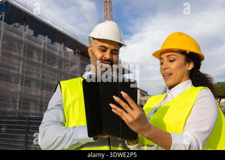Due ingegneri in Hard HAT esaminano i piani di progetto su un tablet in un vivace cantiere edile. Indossano giubbotti riflettenti e si trovano vicino a un par Foto Stock