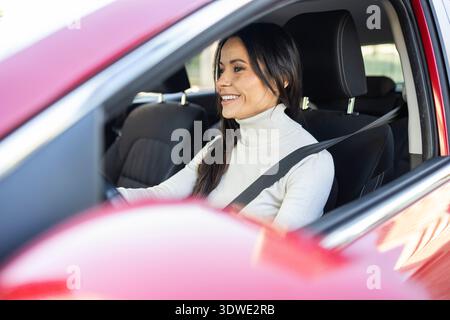 La giovane donna con un maglione bianco sorride mentre guida con fiducia un'auto rossa, godendosi un tranquillo viaggio su strada. Gli interni sono moderni, offrono comfort e Foto Stock