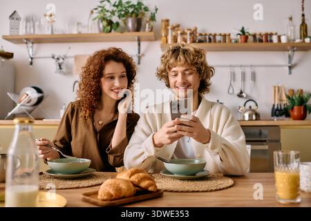 Una coppia felice si diverte a fare colazione, sorridendo mentre guardano il proprio smartphone. Foto Stock