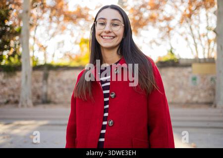 Una donna è vestita calorosamente con un sorprendente cappotto rosso, sorridente mentre ama una fresca giornata invernale all'aperto, con fogliame autunnale sullo sfondo Foto Stock