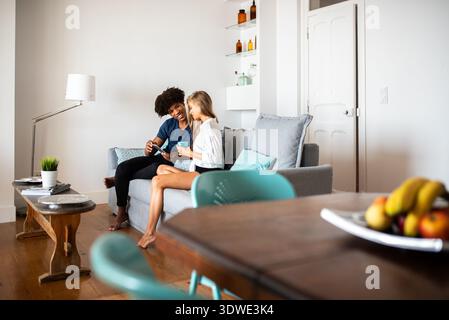Una felice coppia multietnica si rilassa su un comodo divano, condividendo un momento di allegria nella loro casa moderna mentre guardano un telefono insieme, circondati da st Foto Stock