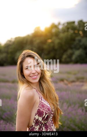 Una giovane donna con i capelli lunghi sorride calorosamente mentre si trova in un lussureggiante campo di lavanda. Il tramonto dorato proietta un bagliore delicato, creando un ambiente tranquillo e pittorico Foto Stock