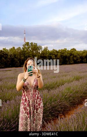 Una donna con un abito a motivi geometrici si trova in un vivace campo di lavanda, catturando la vista panoramica con il suo smartphone. Il paesaggio lussureggiante e gli alberi lontani Foto Stock