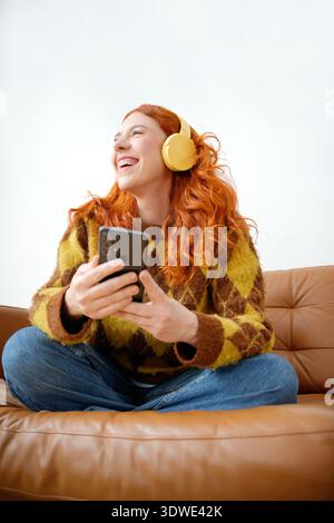 Donna con i capelli rossi che indossa le cuffie gialle, si siede a gambe incrociate su un divano in pelle marrone, sorridendo gioiosamente tenendo lo smartphone in entrambe le mani, e. Foto Stock