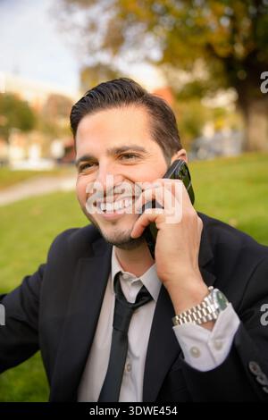 Un uomo d'affari sorridente in tuta parla su uno smartphone mentre è seduto all'aperto in una giornata di sole. Sembra rilassato e felice, incarnando una moderna vita lavorativa Foto Stock