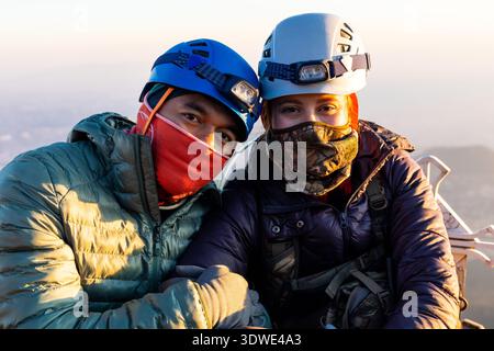 Due escursionisti, raggruppati in giacche e maschere, si godranno un'alba mozzafiato in cima a una vetta della montagna. Il caldo bagliore dell'alba mette in risalto il loro casco Foto Stock