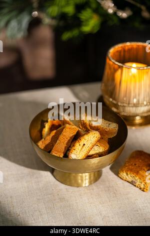 Una ciotola in ottone piena di tradizionali cantuccini italiani alle mandorle. I biscotti sono collocati su una tovaglia luminosa, illuminata da una calda luce di candela, Foto Stock